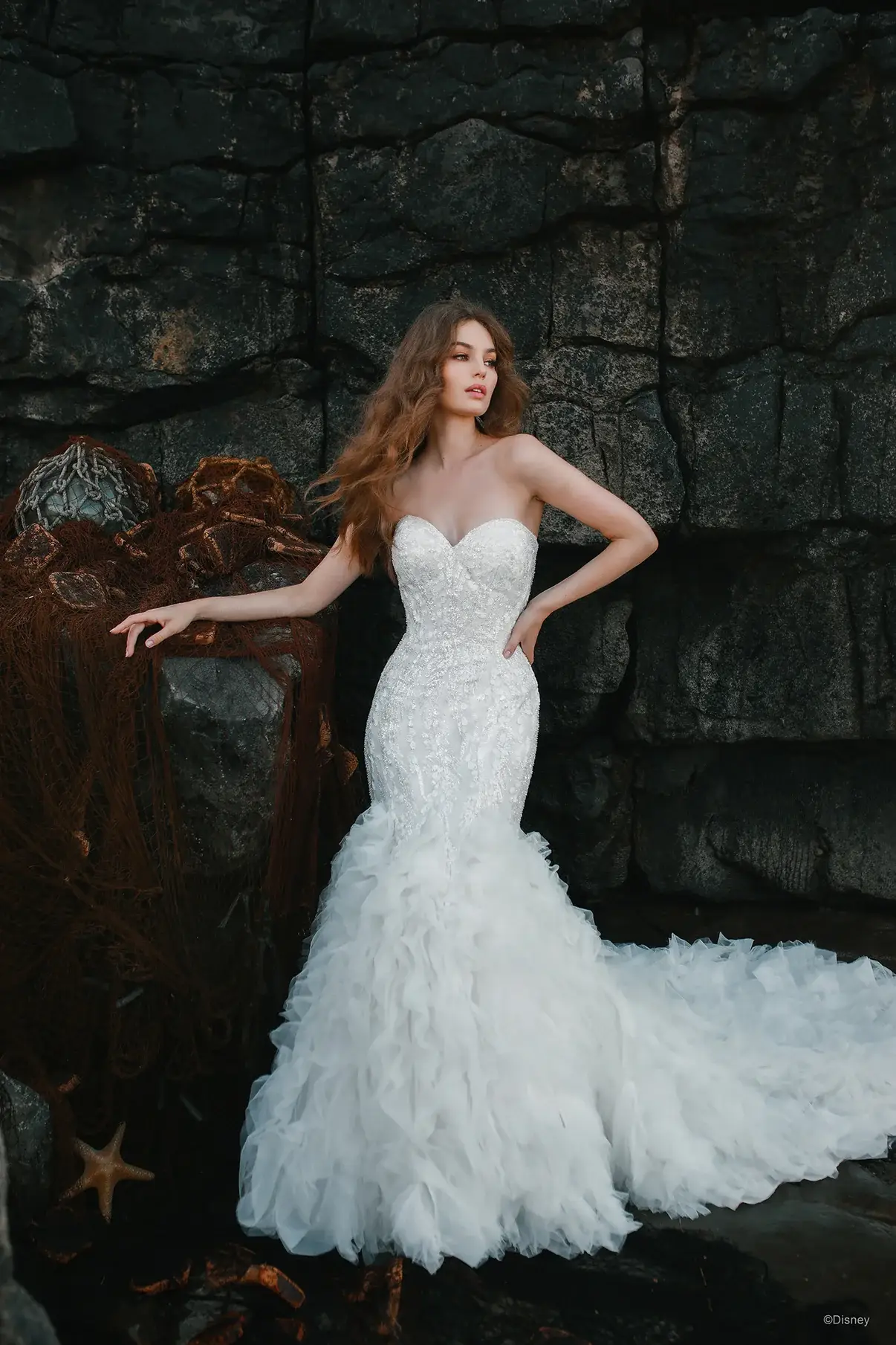 A woman in a strapless, embellished white wedding dress poses against a rocky background. Her hair is wavy and she stands elegantly with one hand on her hip.