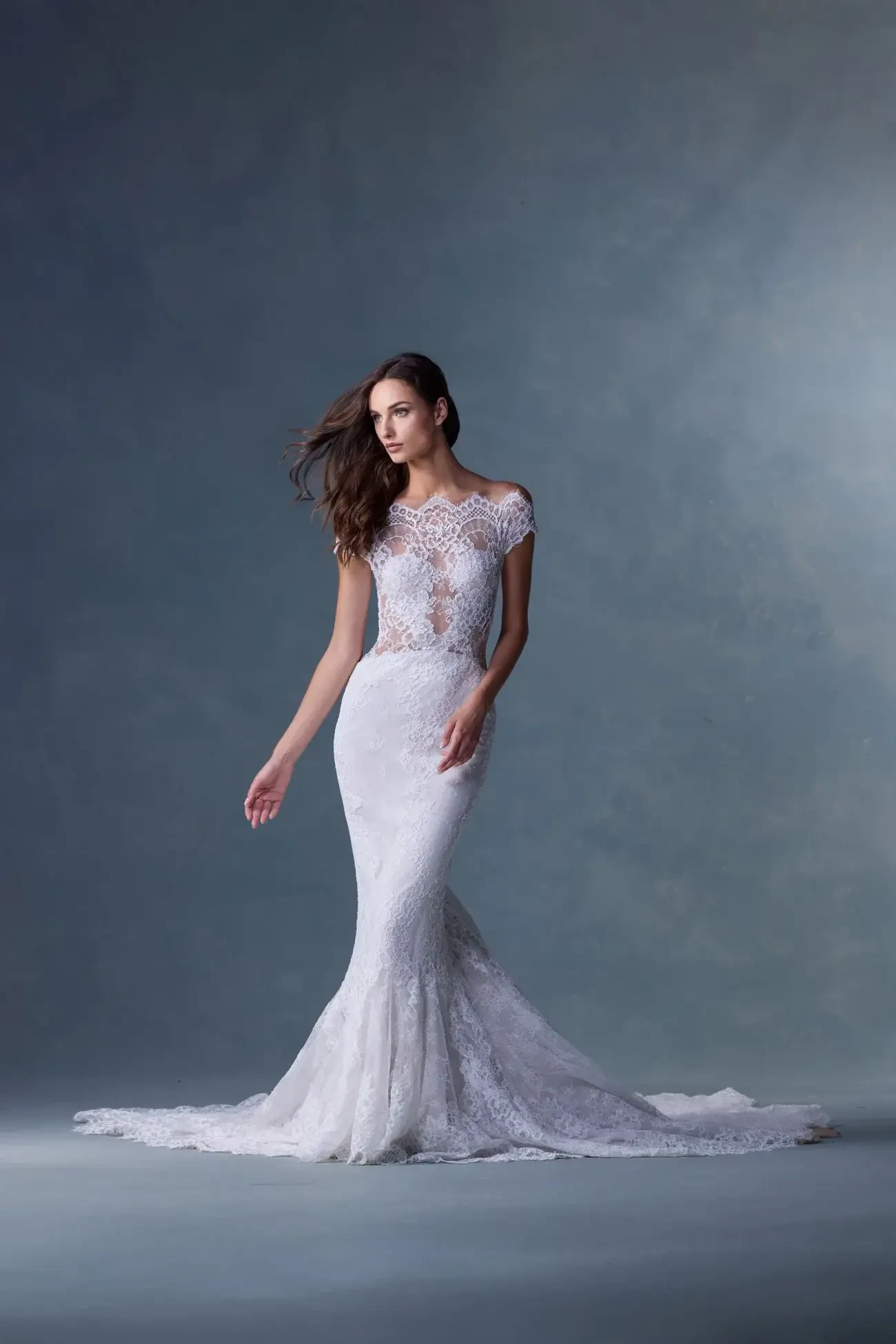 A model wearing a form-fitting, elegant wedding dress with intricate lace details and a sheer bodice, posed against a soft, muted background.