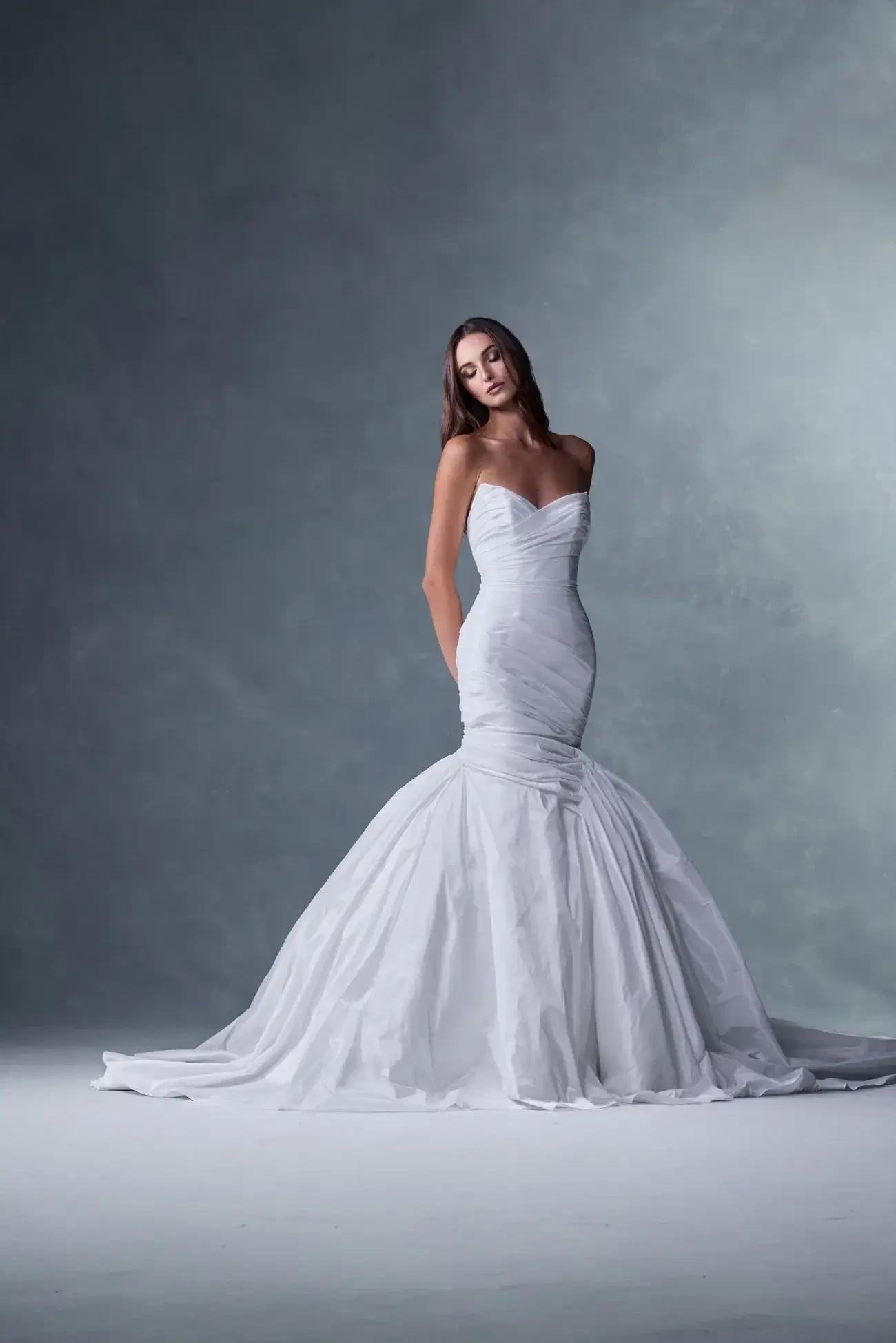 A woman wearing a fitted white wedding gown with a mermaid silhouette, standing against a softly lit backdrop.