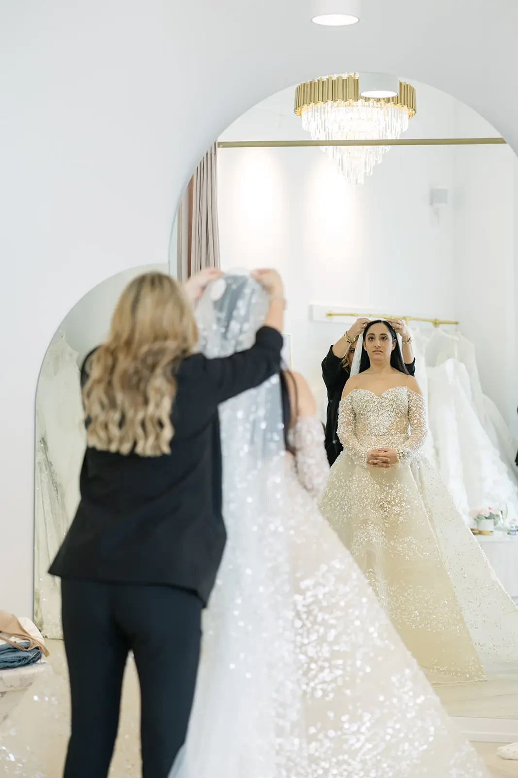 A bride in a sparkling gown stands before a mirror, her expression focused as a stylist adjusts her veil. A luxurious chandelier adds elegance to the scene.