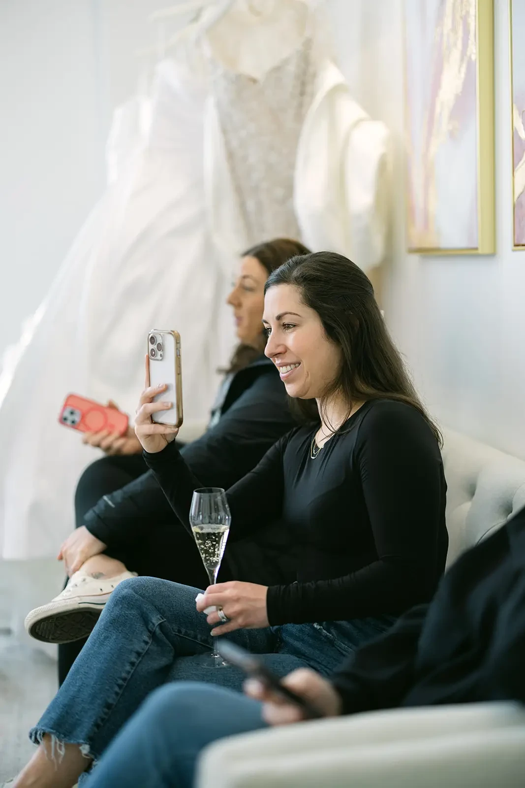 Woman smiling, holding phone for a selfie, with another woman beside her, both seated on a sofa. Champagne glass on lap. Wedding dress hangs in background.