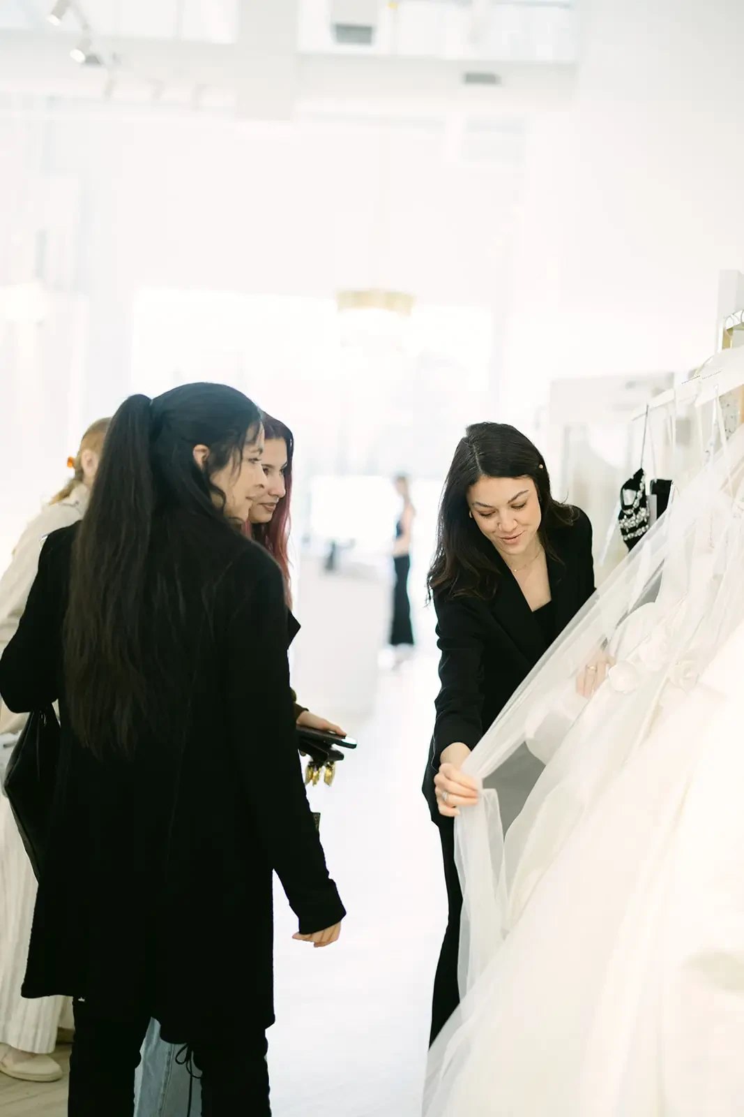 A woman in a bridal shop shows two customers a wedding dress. The setting is bright and elegant, conveying excitement and anticipation.