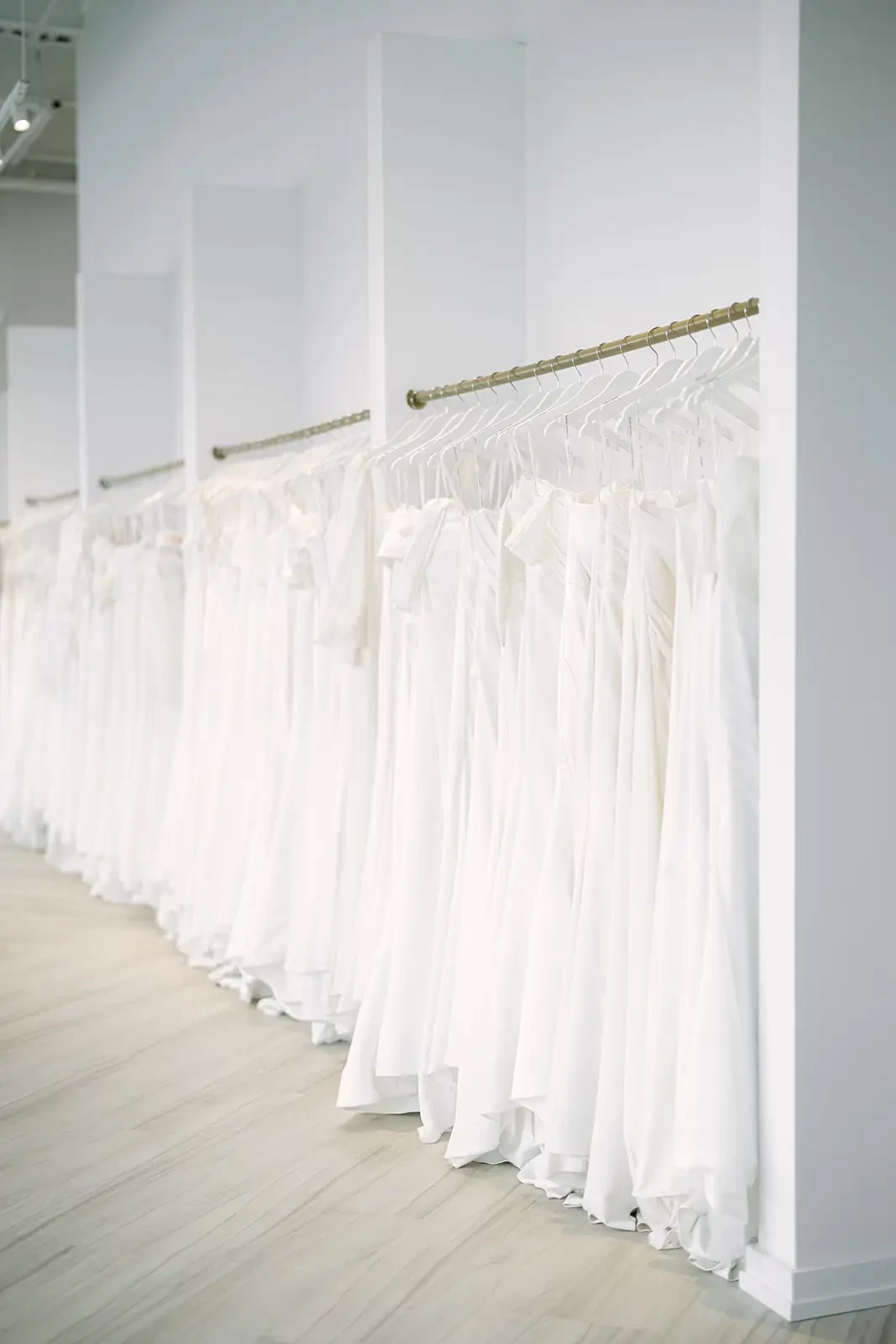A row of white wedding dresses hangs neatly on a rack in a bright, minimalist bridal boutique. The soft lighting and clean space evoke elegance and calm.
