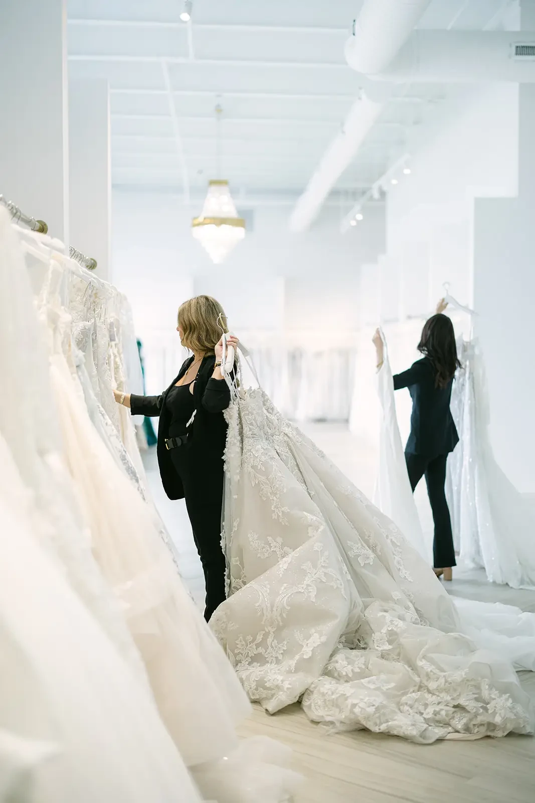 Two women in a bright bridal shop browse through racks of elegant, white wedding dresses. The scene feels serene and meticulous.