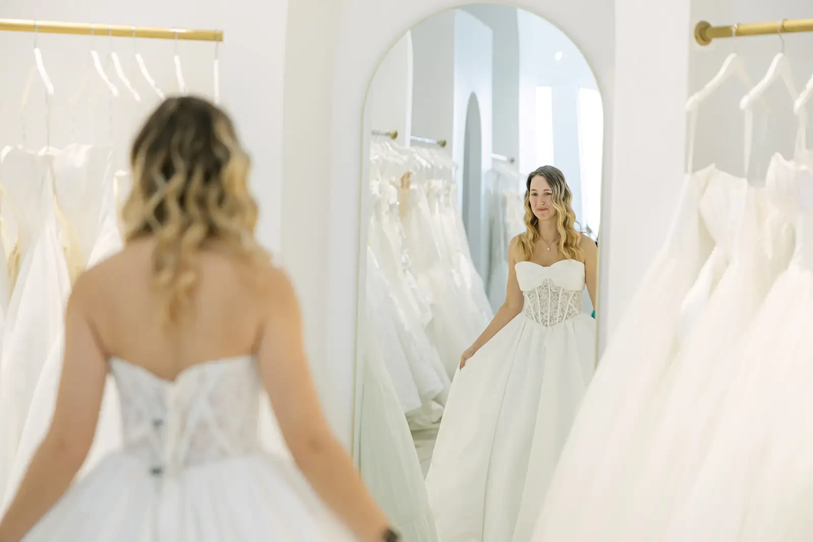 Woman in a bridal boutique smiles at her reflection while trying on a white wedding dress with a corset. Soft lighting and gowns in the background.