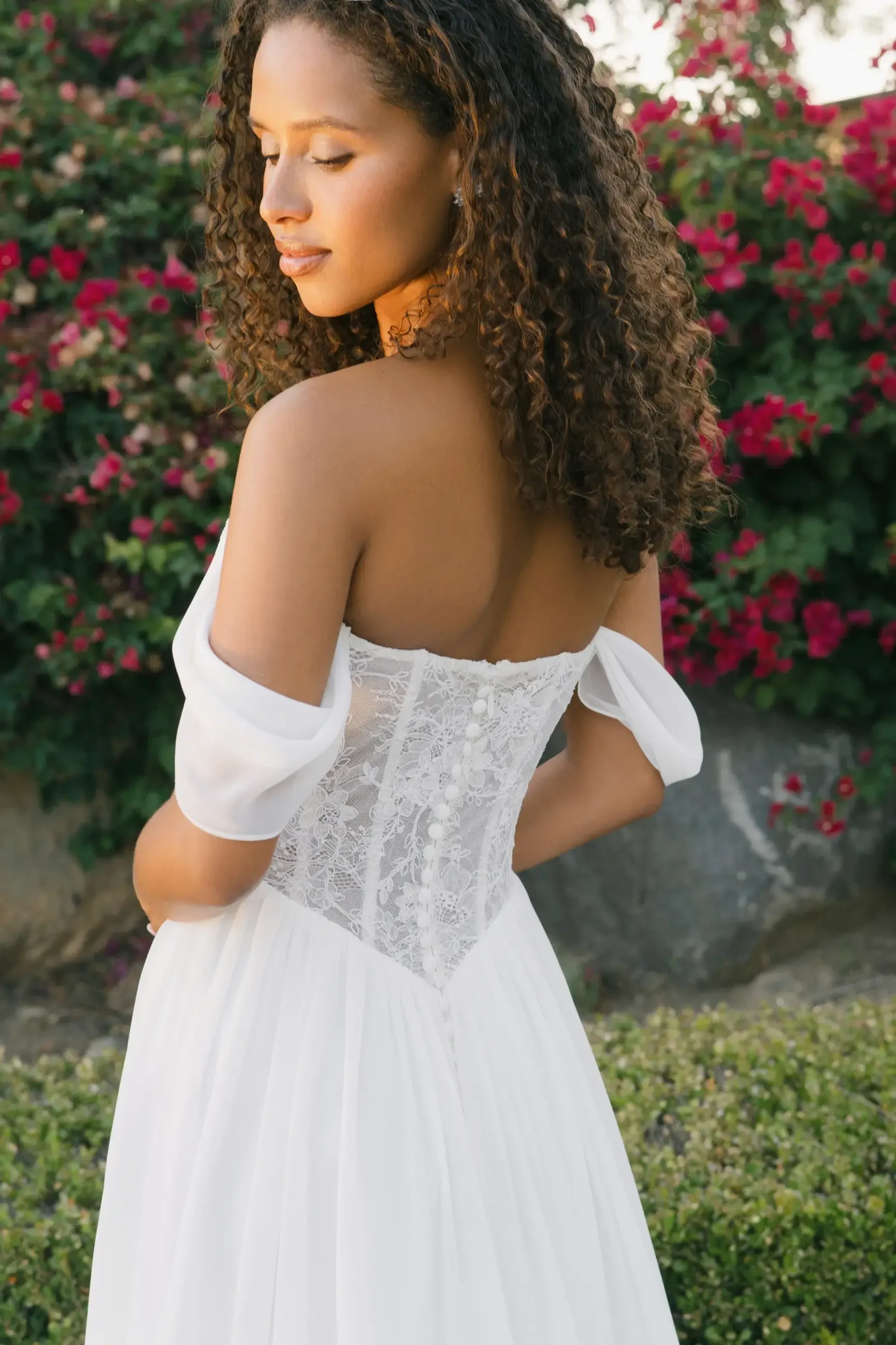 A woman with curly hair is facing away, wearing a white dress with a lace bodice, and surrounded by flowering plants.