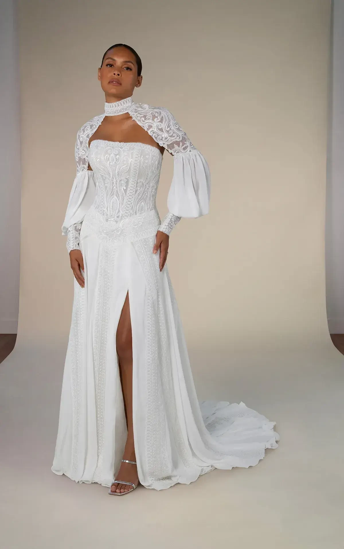 A woman wearing a white gown with long sleeves and a high slit, standing against a neutral background.