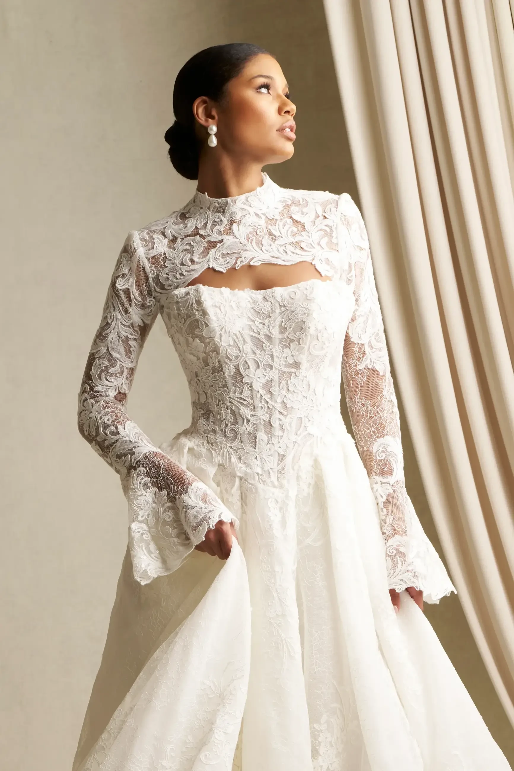 A woman wearing an elegant, long-sleeved lace wedding dress with a high neckline and cut-out detail, standing gracefully near a window with flowing curtains.