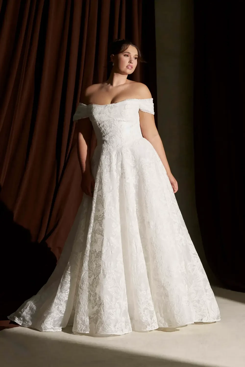 A woman in an elegant, off-the-shoulder white wedding dress standing against a dark curtain backdrop.