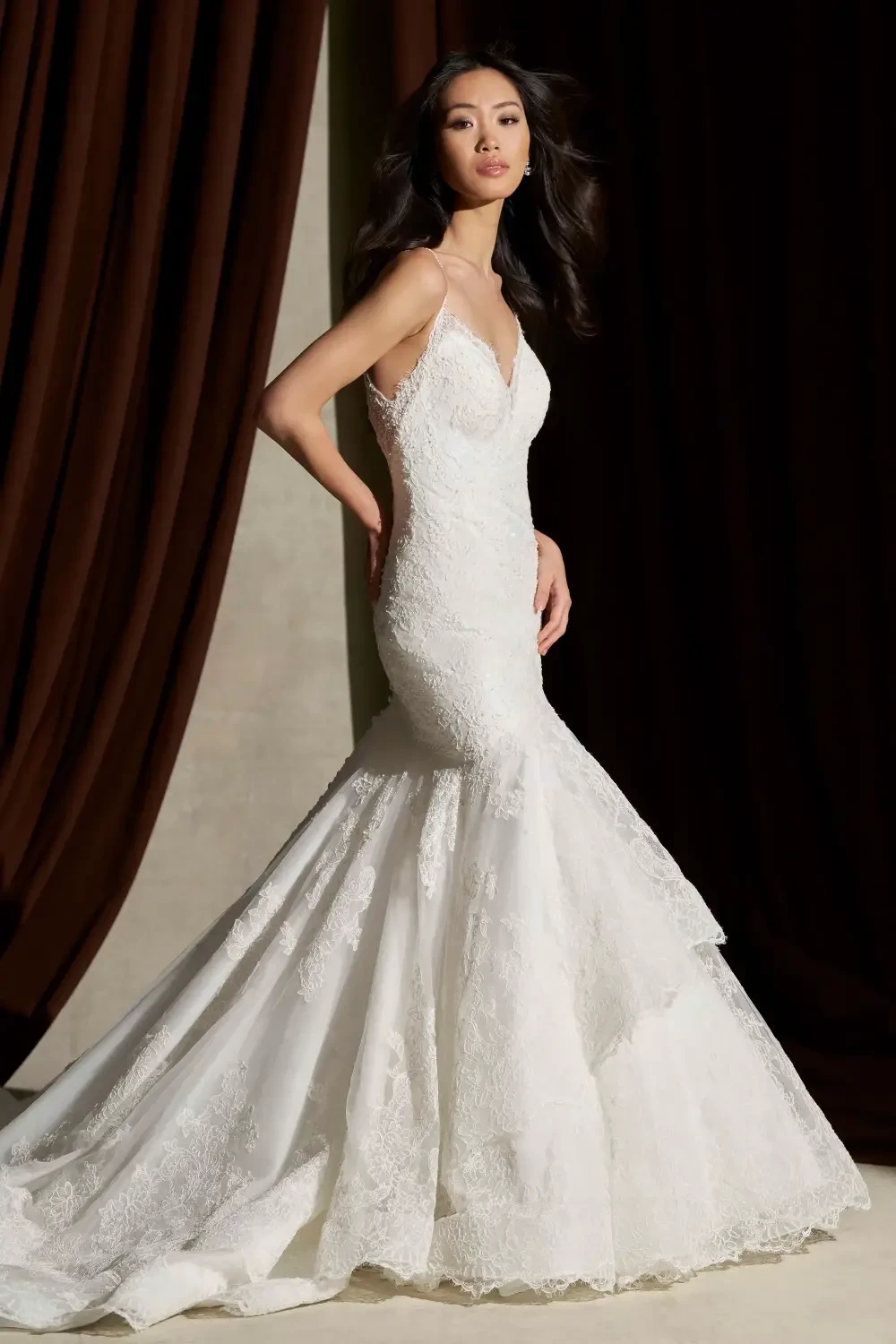 A woman wearing a beautifully designed white wedding dress with intricate lace details, standing elegantly against a dark curtain backdrop.