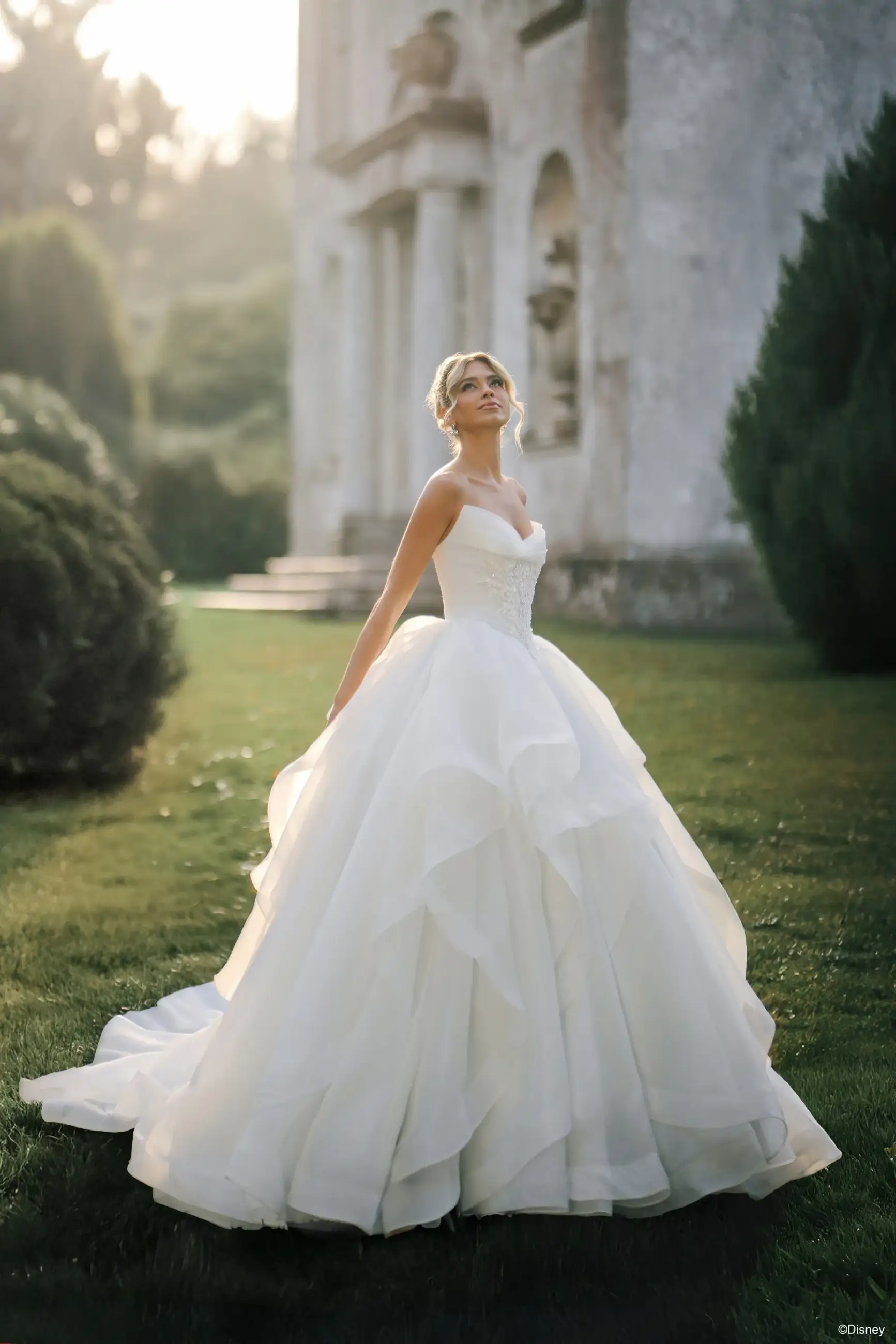 A bride in a large, layered white wedding dress stands gracefully in a garden setting, with trees and an ornate building in the background.