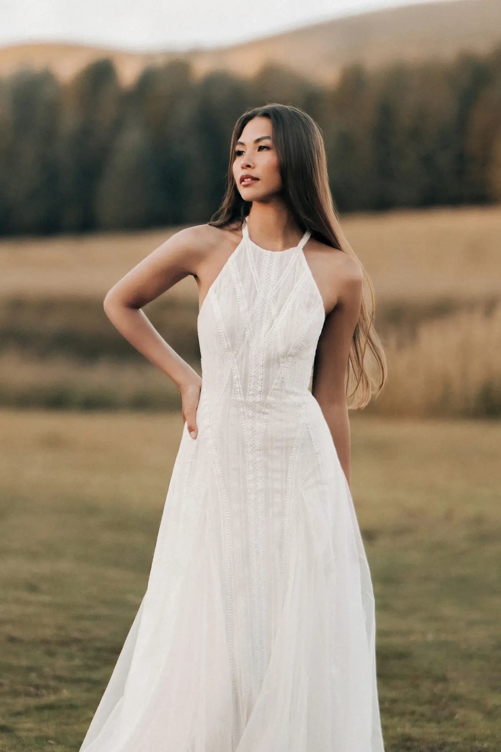 A woman in a flowing white dress stands outdoors, with her hand on her hip and a serene expression. The background features rolling hills and trees.