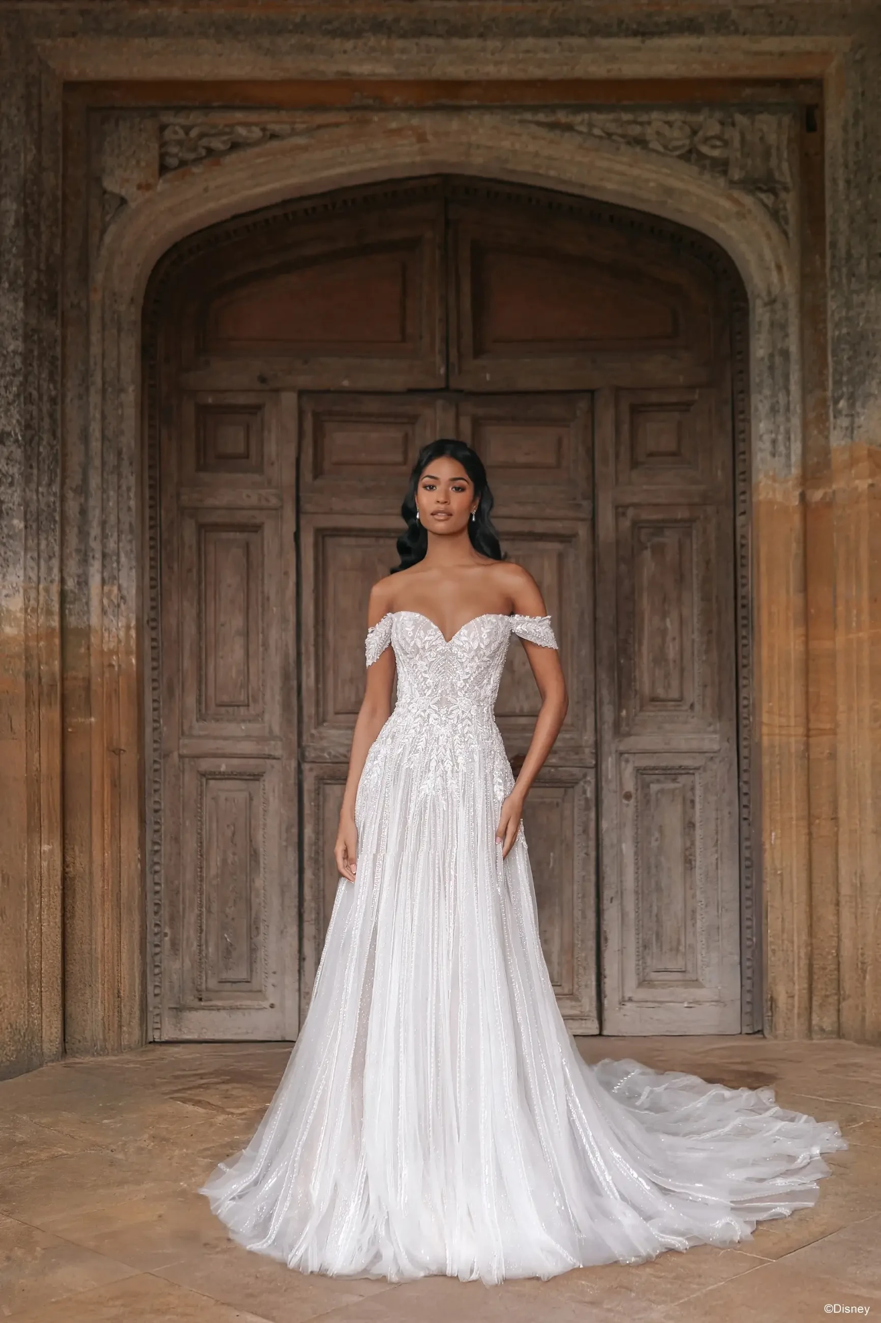 A model in an elegant, off-the-shoulder wedding dress stands in front of a large wooden door with intricate details.