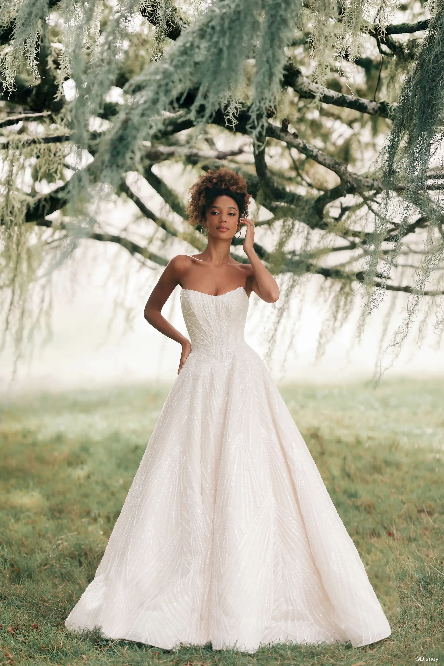 A woman in a strapless, floor-length white gown stands confidently outdoors, surrounded by trees and greenery.
