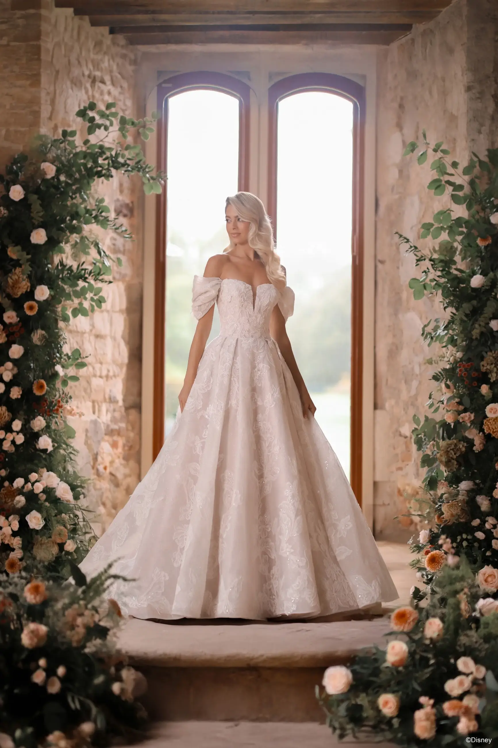 A woman in a formal, off-the-shoulder white gown stands in a beautifully decorated entryway surrounded by floral arrangements. The soft natural light enhances the serene atmosphere of the scene.
