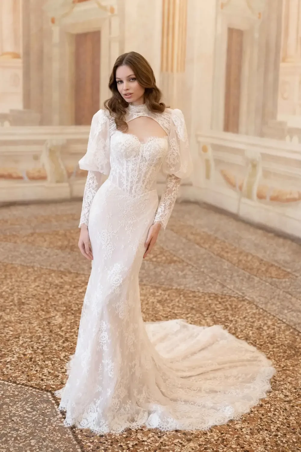 A woman in an elegant white wedding dress with long lace sleeves and a fitted bodice, standing on a decorative floor in a grand setting.