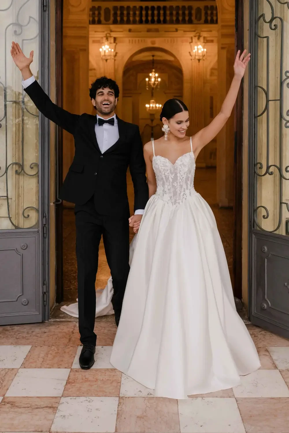 A couple celebrating their wedding outside a grand entrance, with the woman in a beautiful white gown and the man in a black tuxedo, both smiling and raising their arms in joy.
