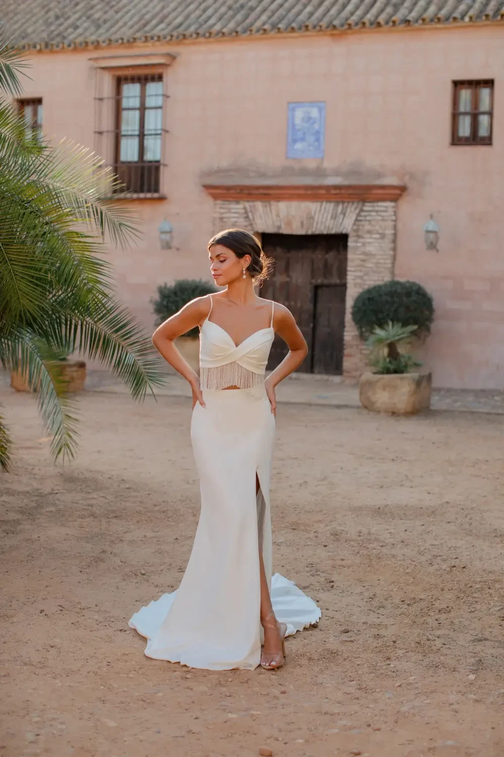 A woman in a white gown stands outdoors, posing confidently with her hands on her hips. The background features a rustic building and greenery.