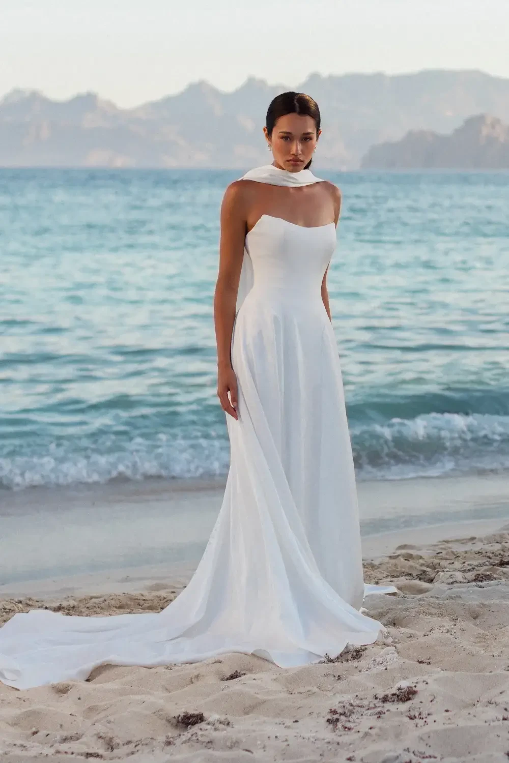 A woman in a white strapless gown stands on a beach with waves and mountains in the background.