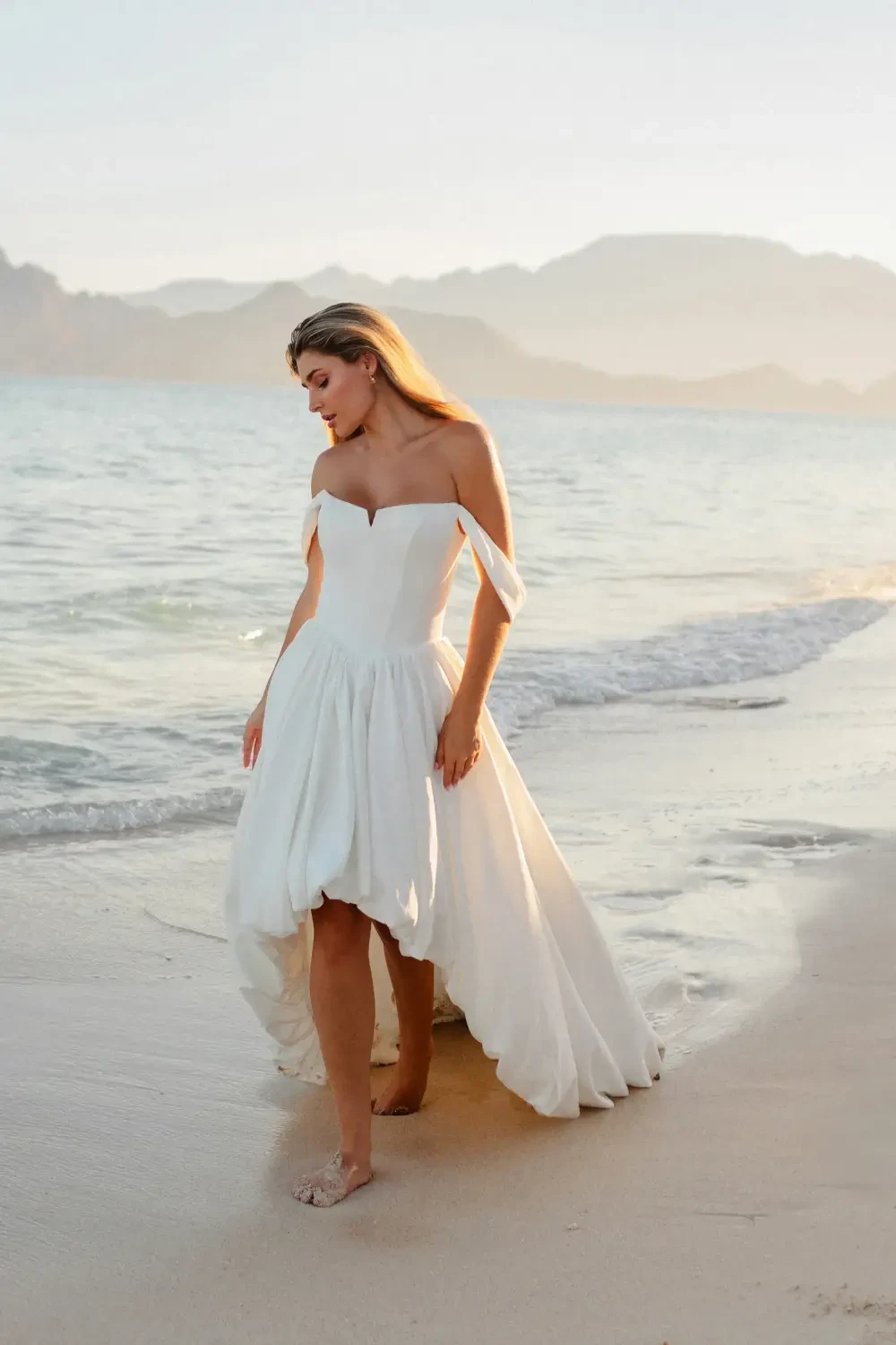 A woman in a strapless white dress walks along the beach near the water with mountains in the background.