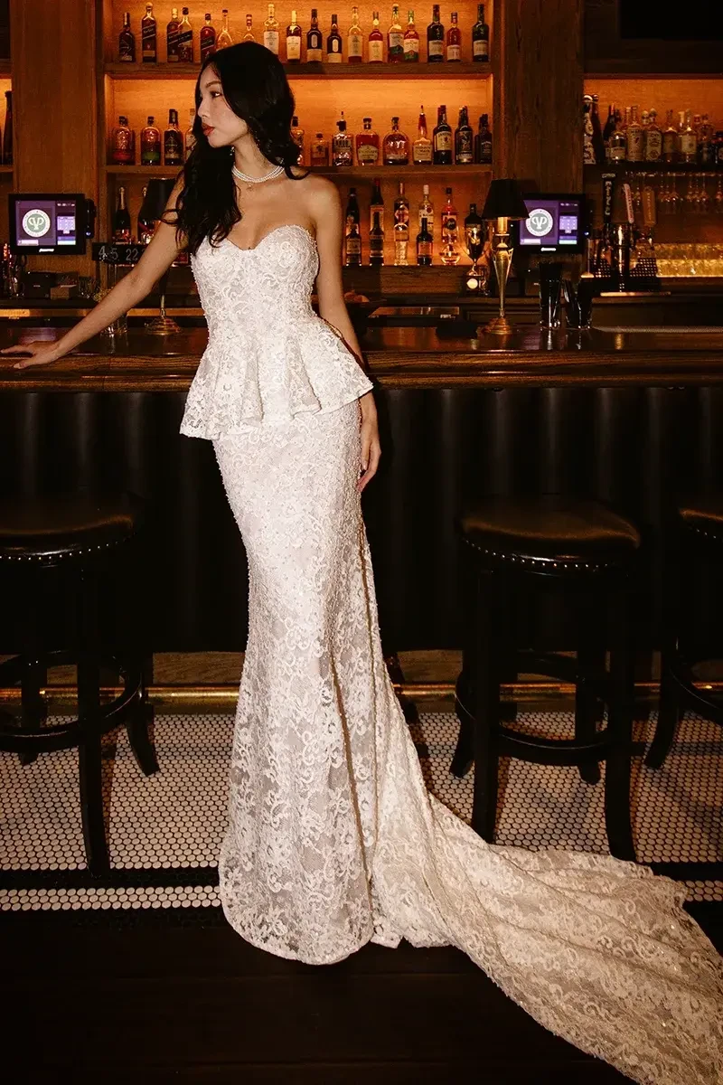 A woman stands elegantly at a bar, wearing a strapless white lace gown with a peplum design and a long train.