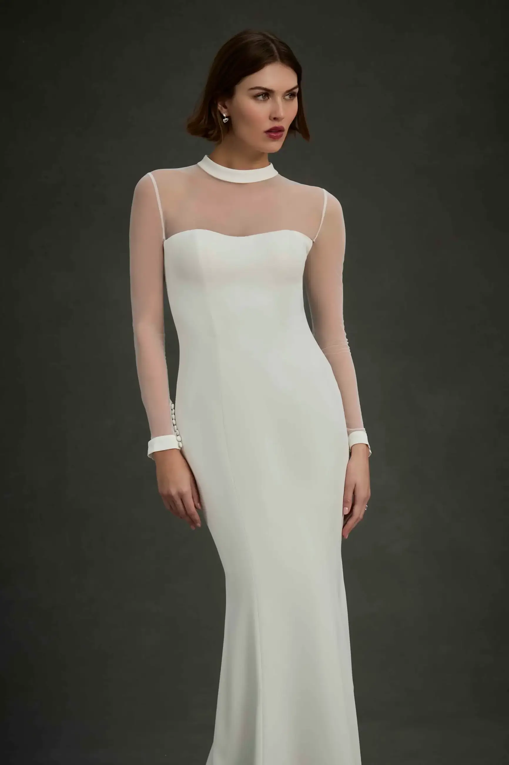 A model wearing a sleek, white evening gown with a fitted silhouette and sheer long sleeves, set against a dark background.
