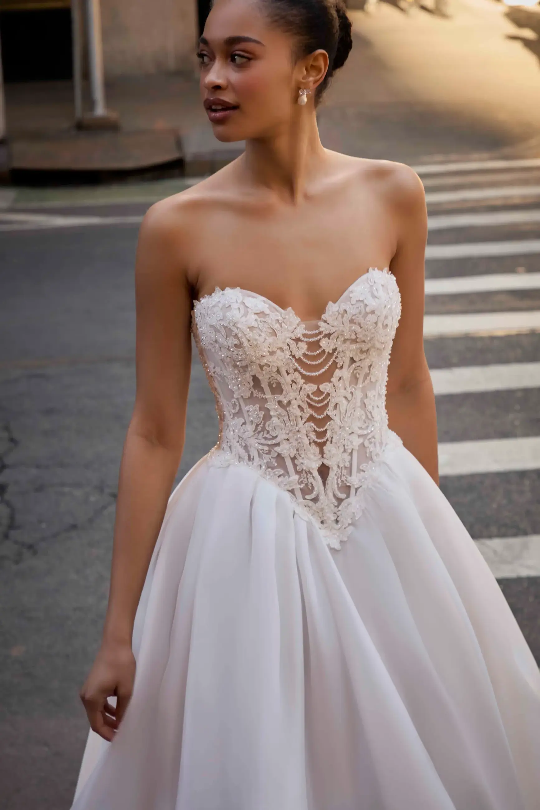A woman wearing a strapless wedding dress with intricate lace detailing on the bodice and a flowing skirt, standing at a crosswalk.