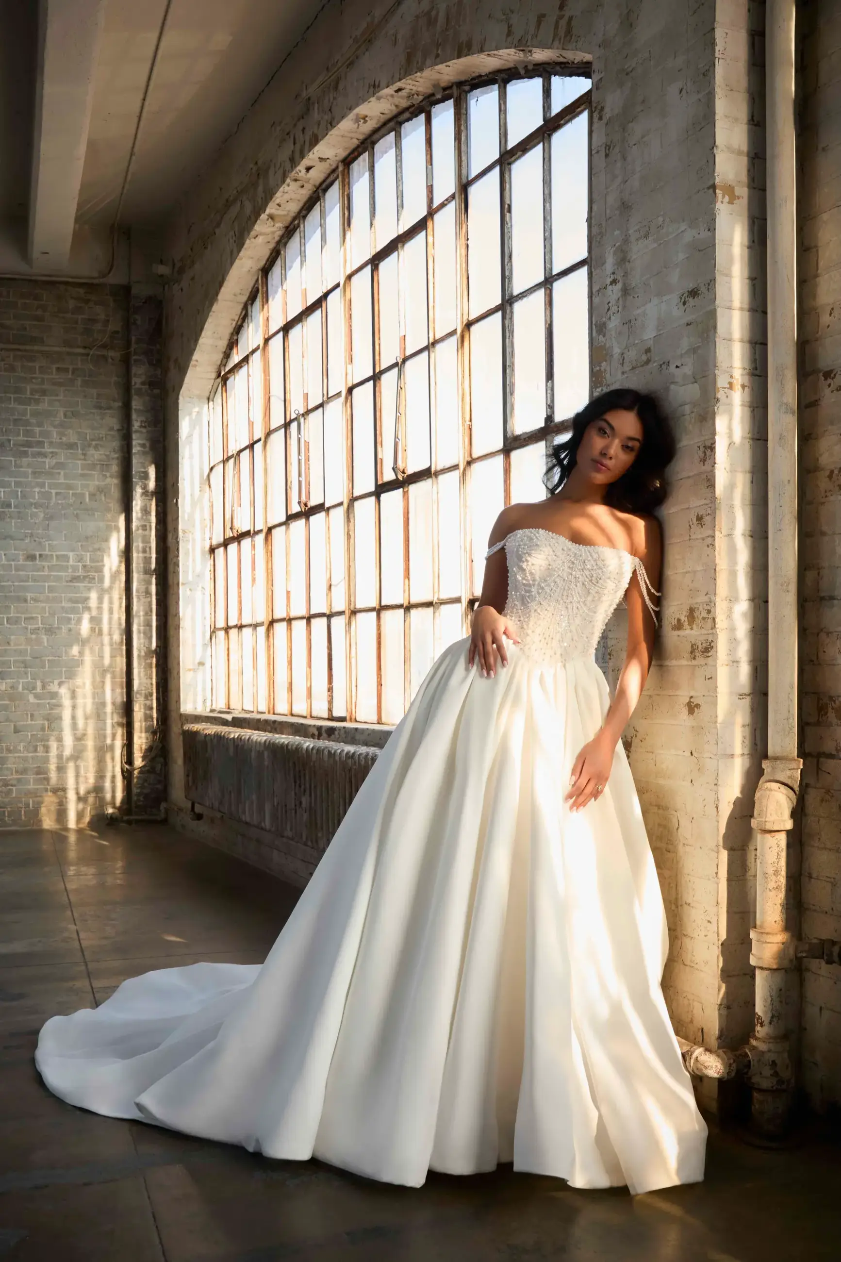A woman in a strapless white gown leaning against a large window with natural light streaming in.