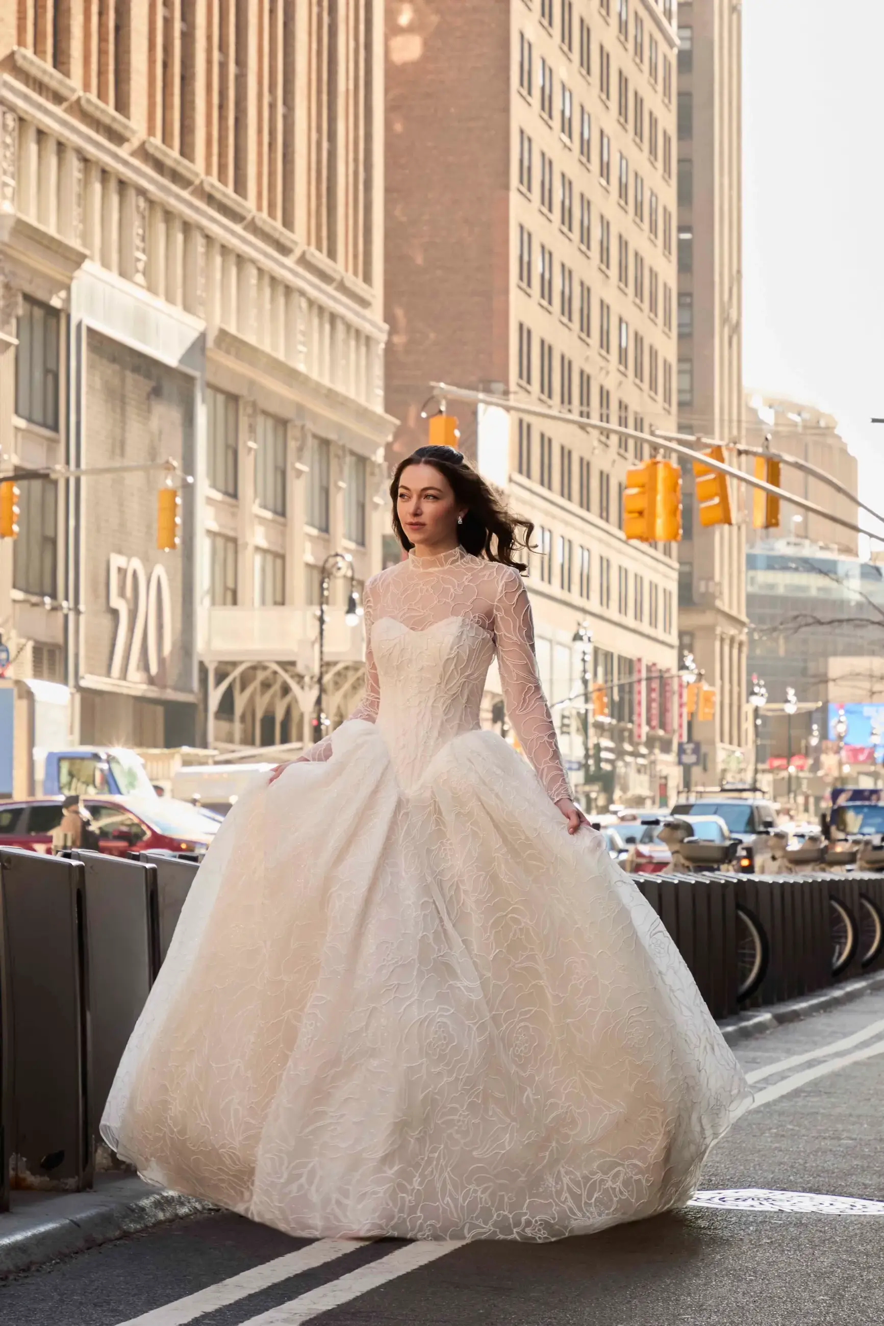 A woman in a luxurious white wedding dress is walking confidently down a city street, surrounded by tall buildings and traffic. The dress features intricate lace details and has a voluminous skirt.
