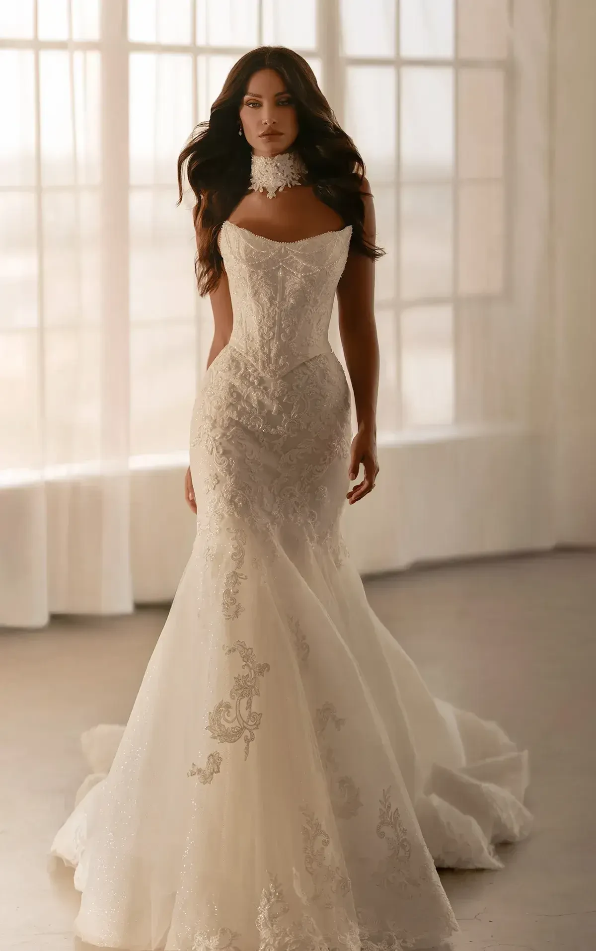 A woman wearing an elegant wedding dress with intricate lace details, standing in a sunlit room with large windows. Her hair is styled in loose waves, and she has a choker around her neck.
