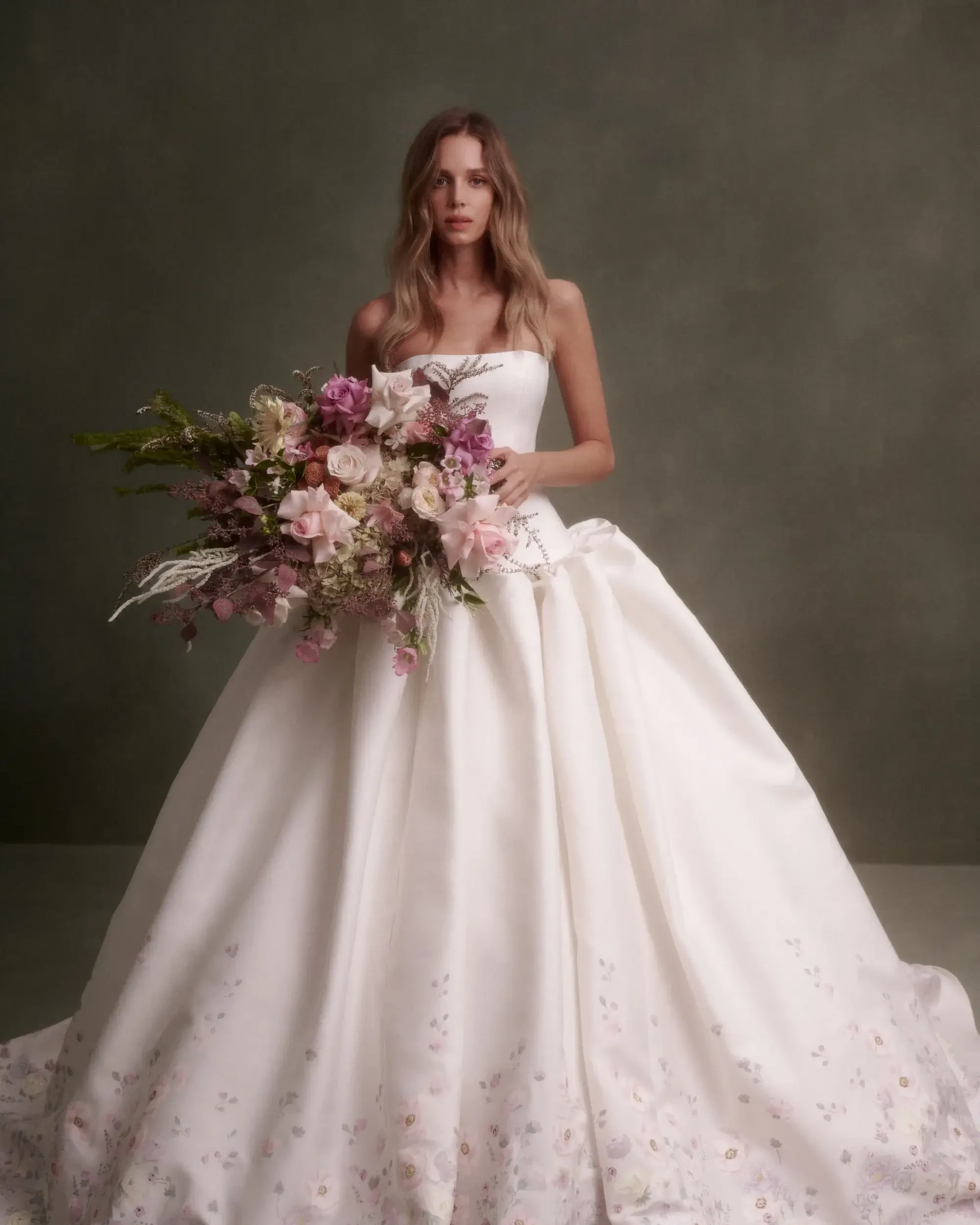 A model wearing a strapless white wedding dress, holding a bouquet of flowers, in front of a muted green background.