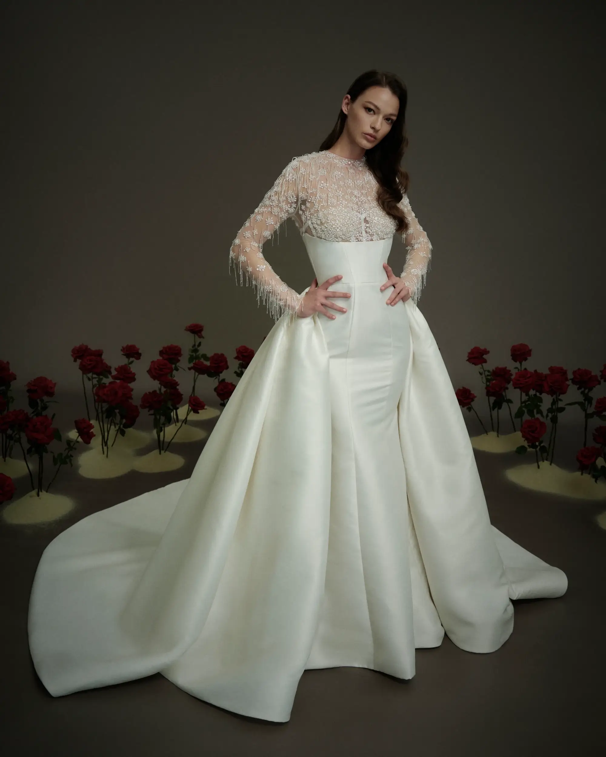 A model wearing an elegant white gown with a fitted silhouette and dramatic train, complemented by a sheer, beaded long-sleeve top. The background features red roses on pedestals.