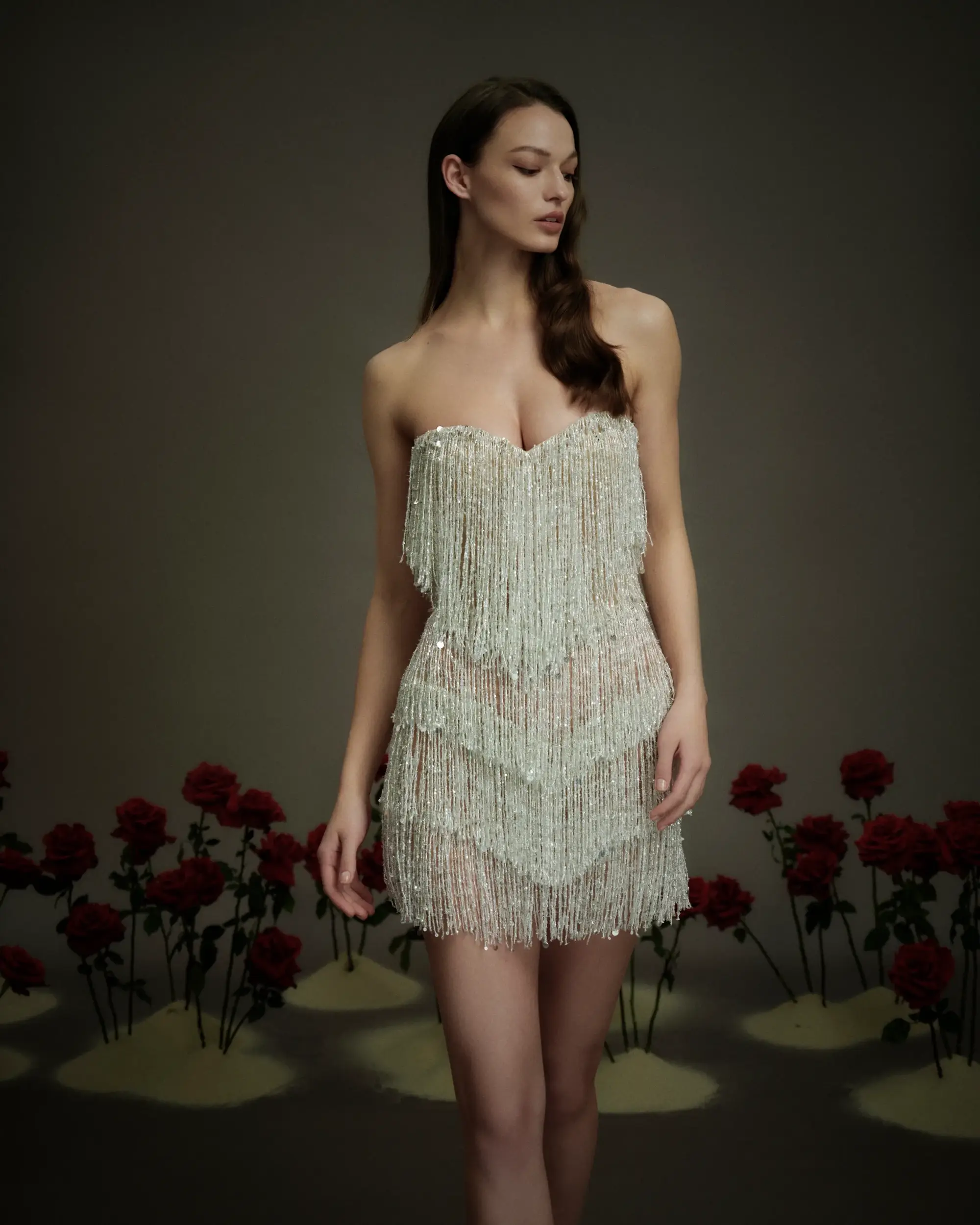 A model wearing a sparkling, fringed dress stands gracefully in front of a backdrop of roses.