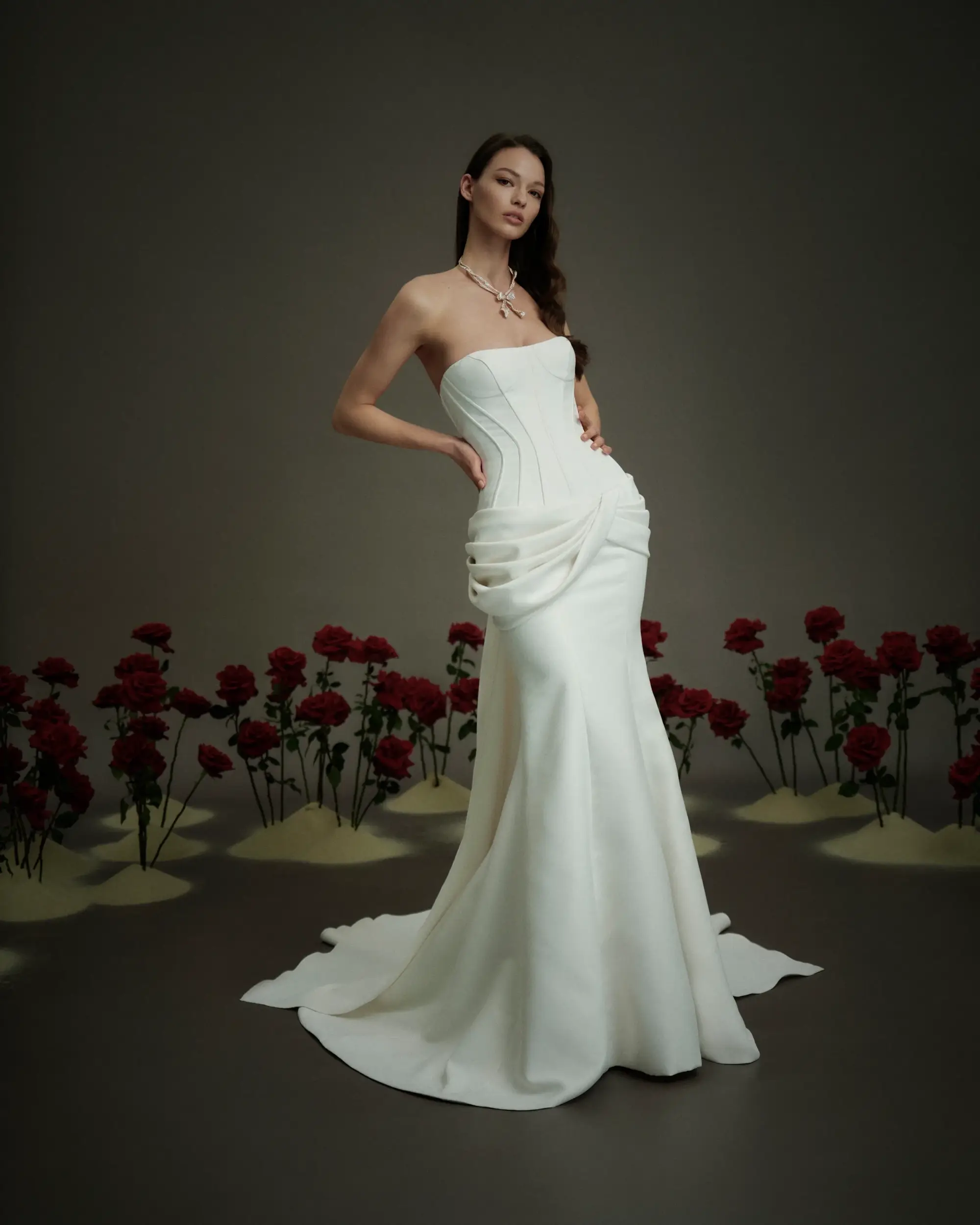 A model poses in a strapless white gown with ruched detailing, surrounded by red roses on a sandy backdrop.
