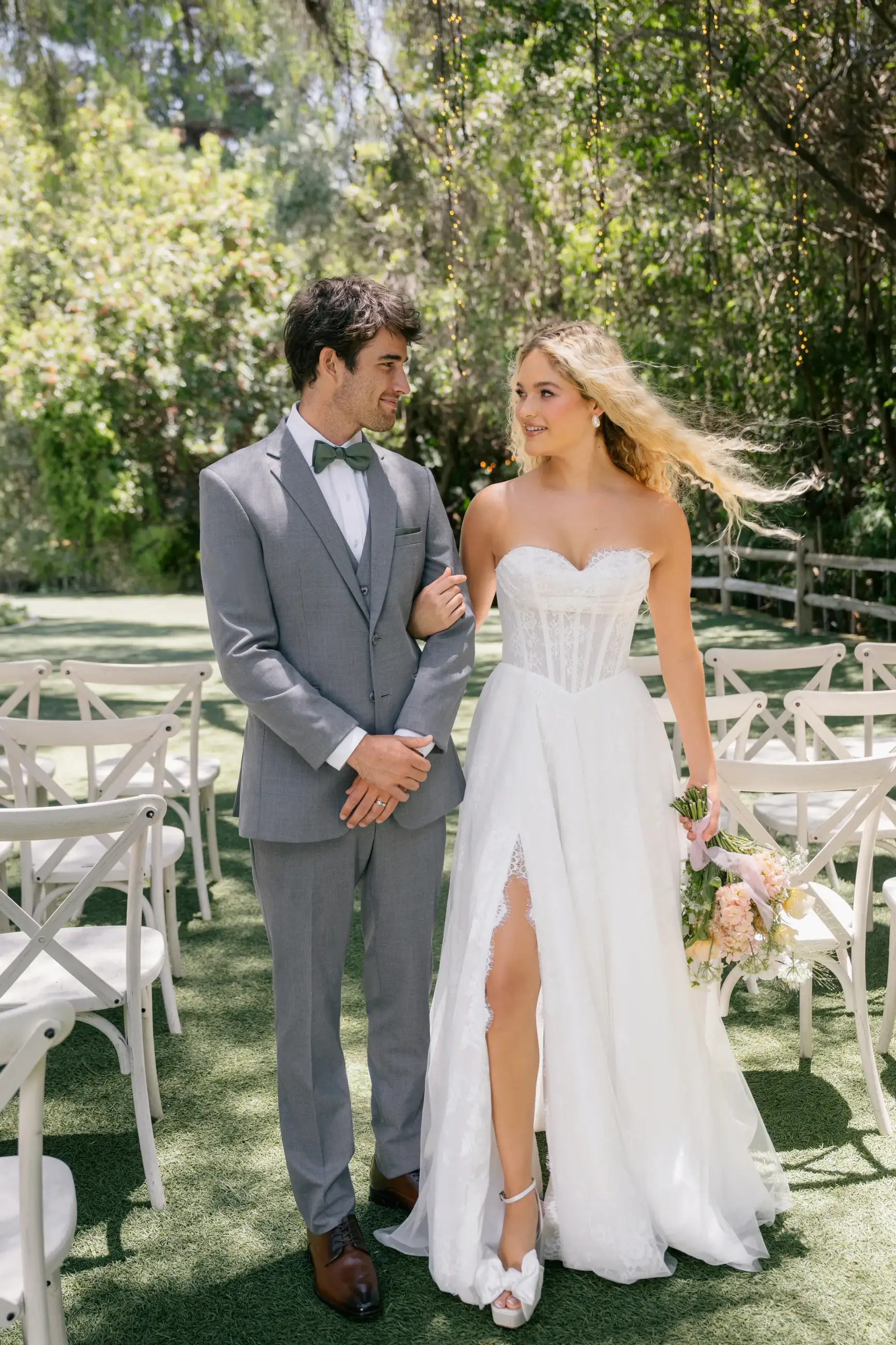 A couple dressed in formal wedding attire stands together outdoors, smiling at each other. The bride is in a white gown with a thigh-high slit and holds a bouquet of flowers, while the groom wears a gray suit with a bow tie. The setting features greenery and chairs arranged for a ceremony.
