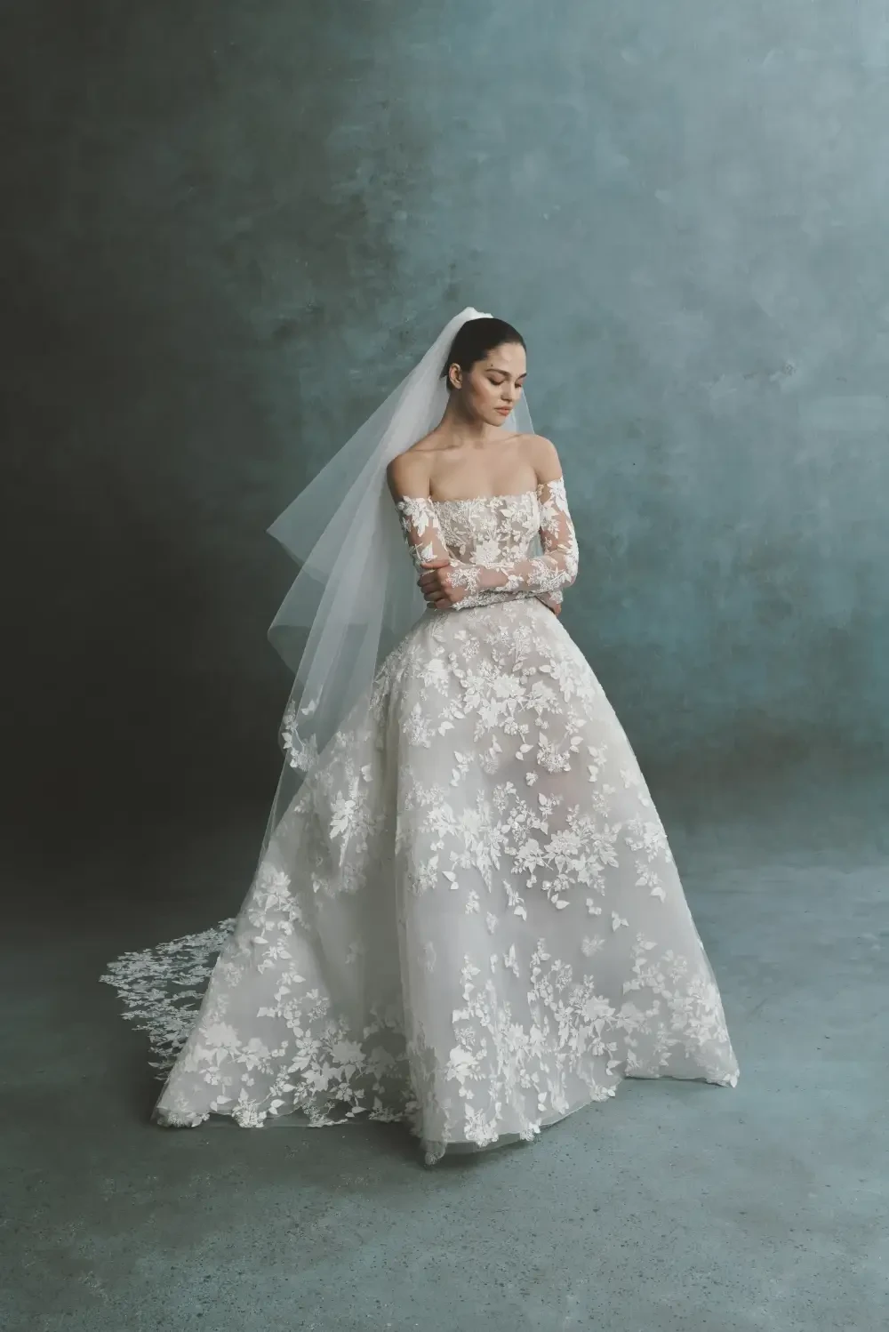 A bride in an elegant off-the-shoulder wedding dress with intricate lace detailing, wearing a long veil, standing against a textured blue background.