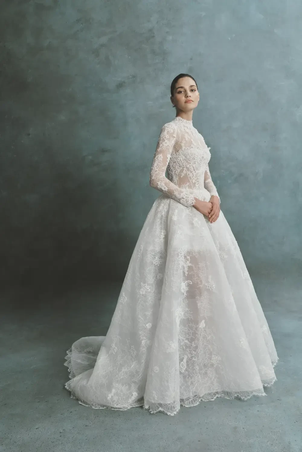 A model wearing a long-sleeved, intricately designed white lace wedding dress, standing gracefully against a textured backdrop.