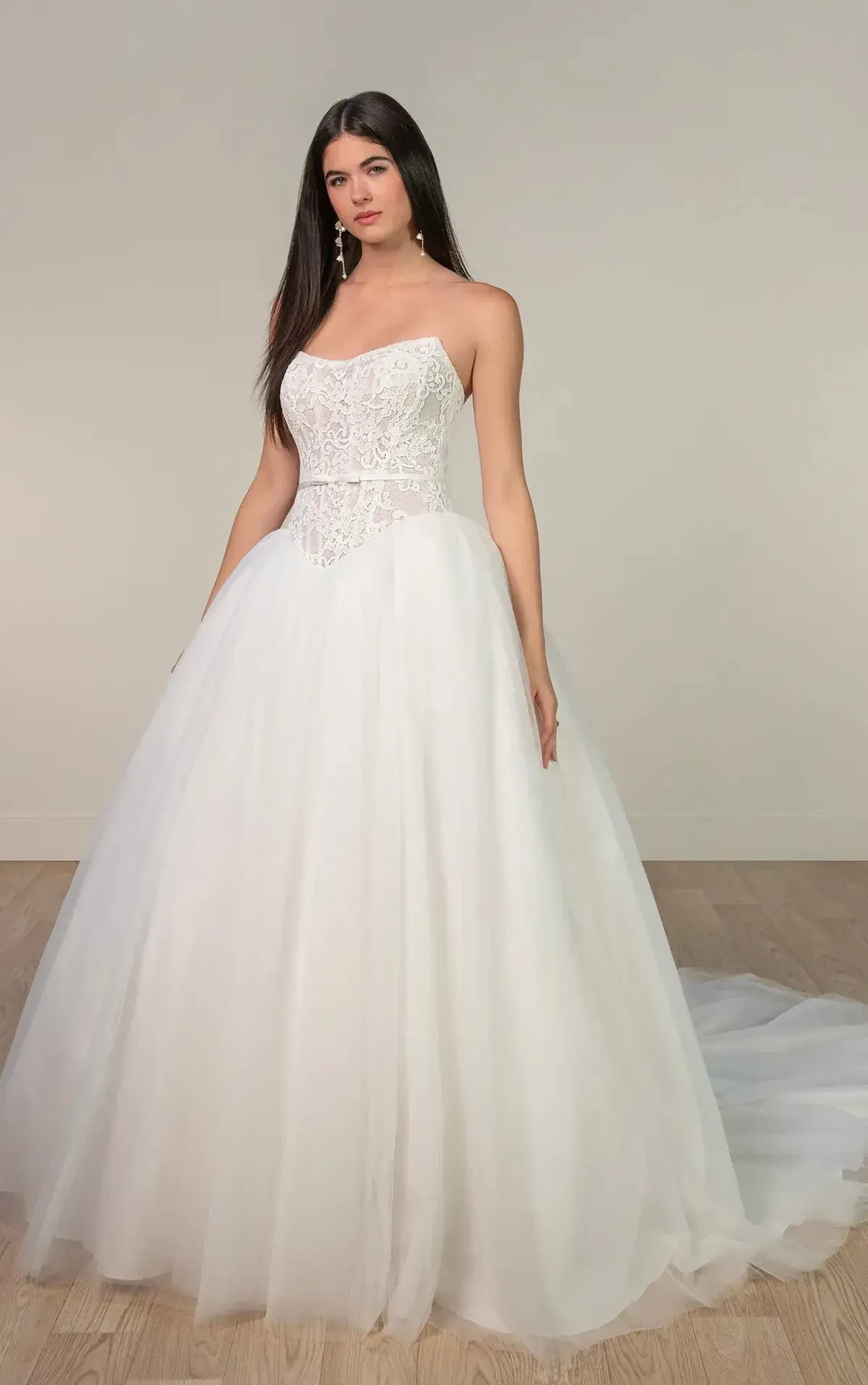 A model wearing a strapless, white wedding gown with a fitted bodice and a full tulle skirt. The dress features intricate beading and embroidery on the bodice.
