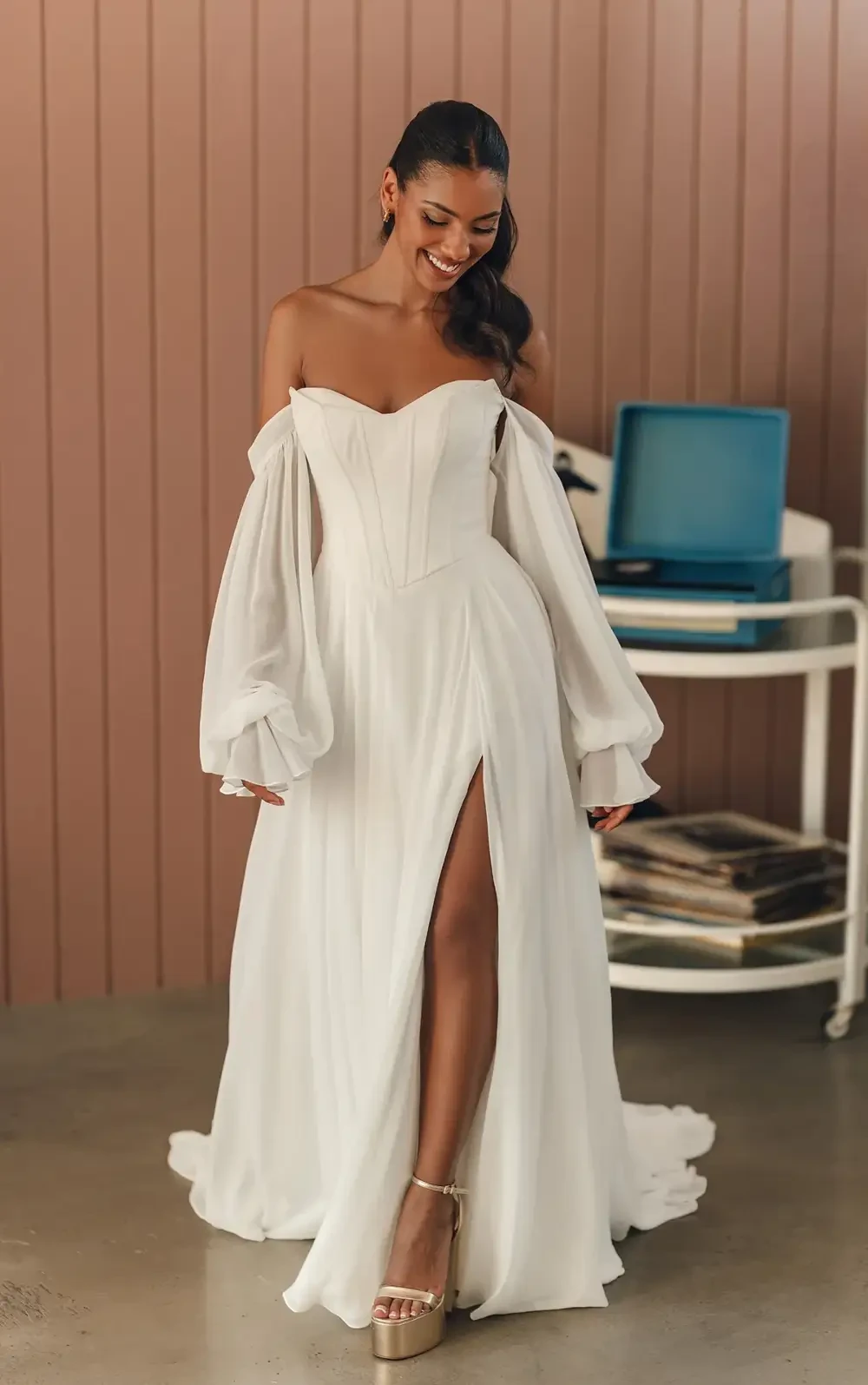 A woman in a white off-shoulder gown with a high slit, standing in a room with a vintage record player and bookshelf in the background.