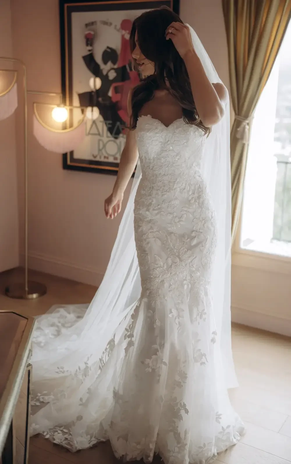 A bride in a beautiful lace wedding dress with a fitted bodice and a flared skirt, holding her veil, standing in a well-lit room with elegant decor.