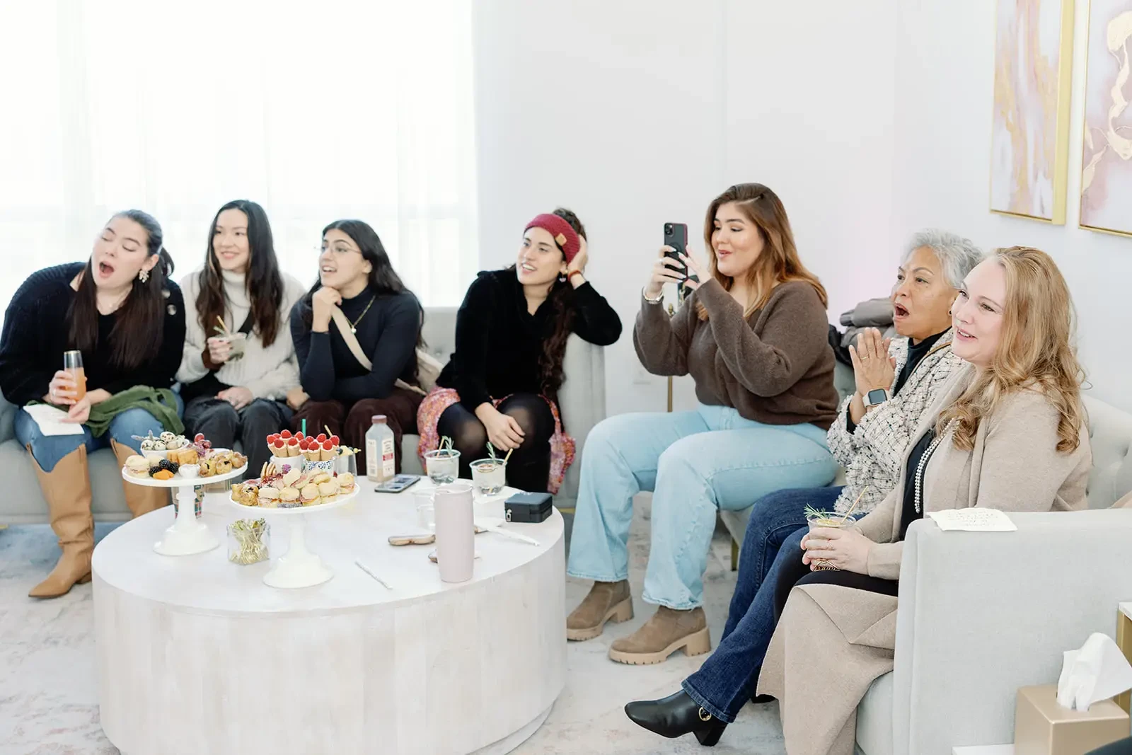A group of women sitting together in a bright, stylish room, enjoying snacks and drinks. They appear to be engaged in a lively conversation or activity, with some laughing and others taking photos.