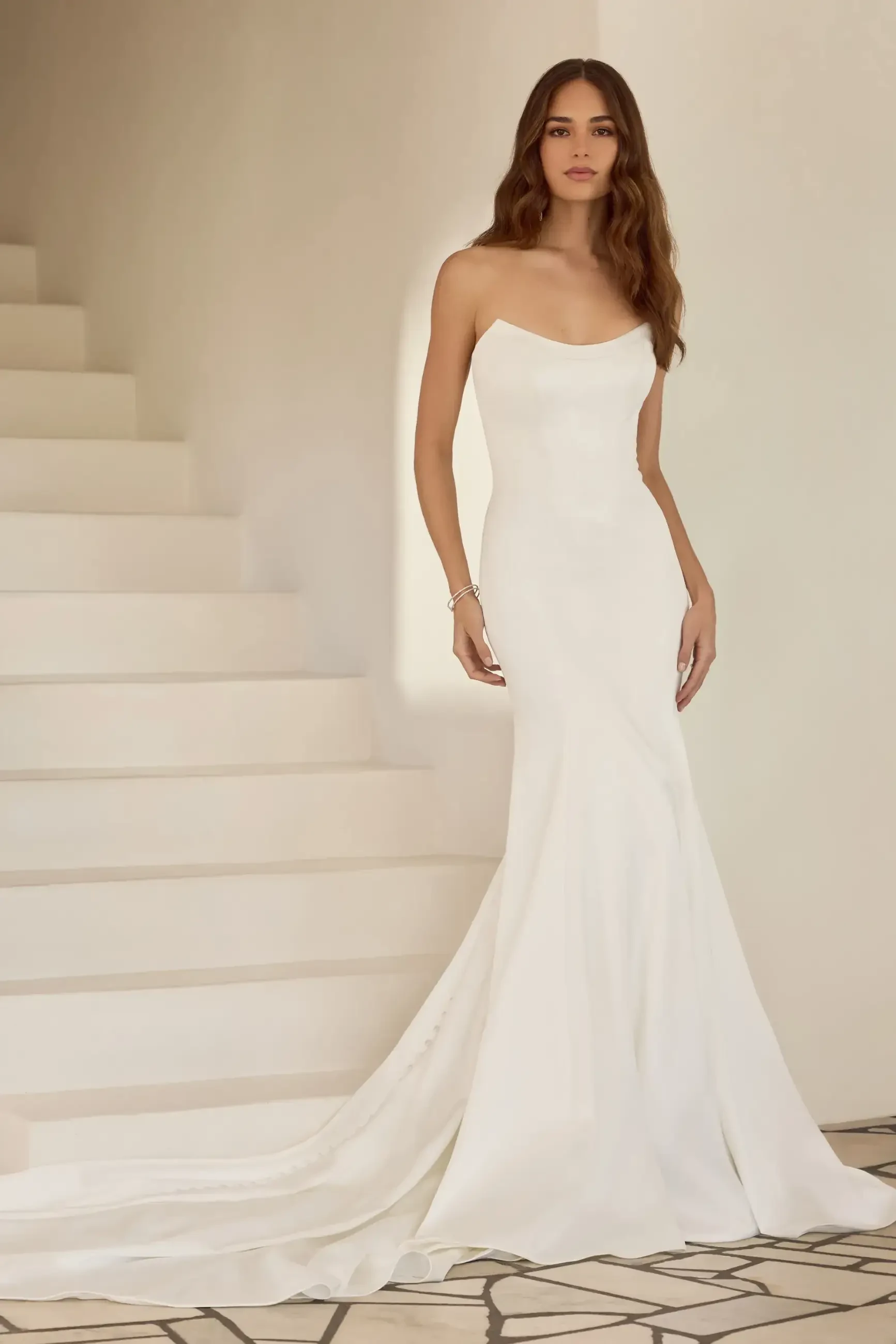 A woman wearing a fitted white gown with a slight train, standing on a staircase. The gown has one shoulder strap and the fabric appears smooth and elegant.